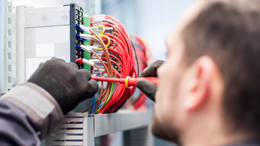 Électricien ganté effectuant la maintenance du câblage électrique avec un tournevis dans une armoire technique.